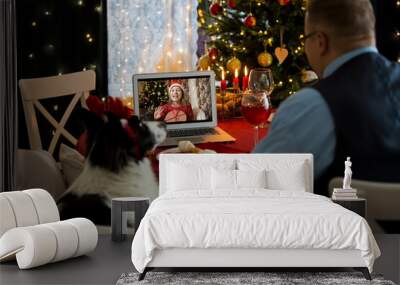 Mature man celebrating Christmas with his dog sitting at served holiday  table with laptop. People greeting their friends on video call using webcam. Christmas eve online. New normal social distancing Wall mural