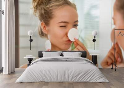 Young beautiful woman cleaning her face skin with cotton pad in bathroom. Standing in towel, looking in the mirror, laughing and having fun. Morning skincare routine.   Wall mural