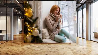 Young beautiful woman with reddish hair sitting home by the window with cup of hot coffee wearing knitted warm sweater. Christmas tree with decorations and lights in the room, snowy winter outside Wall mural