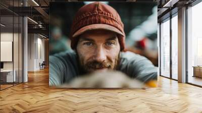 A bearded man in a red hat and casual attire is smiling and looking directly at the camera, creating an approachable and friendly atmosphere in a bustling environment. Wall mural