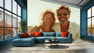 A joyful couple with windswept hair, wearing sunglasses and casual attire, enjoying a sunny day outdoors with a vibrant blue sky and sandy surroundings. Wall mural