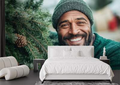 A joyful man wearing a green knit cap shows a bright smile next to a lush green wreath with pinecones, capturing the essence of holiday spirit in the park. Wall mural