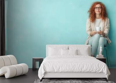 A red-haired woman in glasses and casual attire, sits relaxed with a cup in her hands against a vibrant teal backdrop, exuding calm and stylish ease. Wall mural