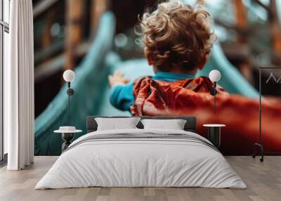 A young child with curly hair and a cape reaches towards the top of a slide in an outdoor playground, showcasing a playful and adventurous moment in motion. Wall mural