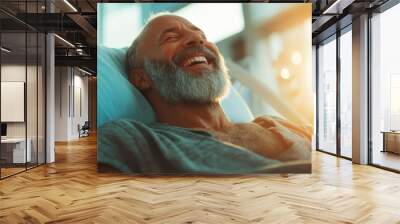 An elderly man with a beard lies in a hospital bed, laughing joyfully in a sunlit room, capturing a moment of happiness and relief in a healthcare setting. Wall mural