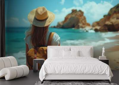 Back view of a woman with a straw hat and backpack looking out at the clear blue sea and rocky coastline Wall mural