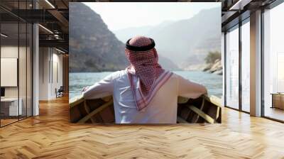 A Saudi Arabian man gazes into the tranquil river, surrounded by majestic mountains on a serene journey at dawn Wall mural