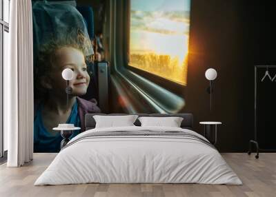 Beautiful little girl looking out train window outside, while it moving. Going on vacations and traveling by railway in summer. Sunset time. Wall mural