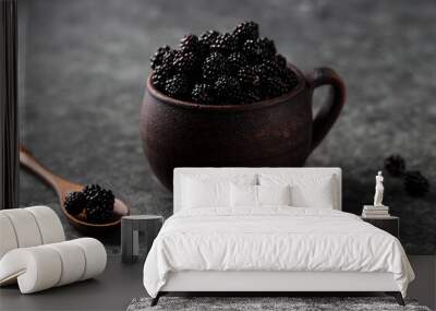 Blackberries in an earthenware mug are piled with a hill. Next to it is a wooden spoon in which the berries are scattered around on the table Wall mural