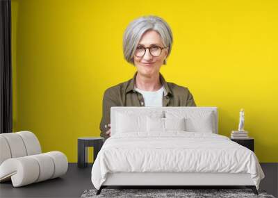 Charming smiling mature woman in her 50s in eye glasses standing with folded hands wearing green shirt with white t-shirt isolated on yellow background. Mature people beauty concept. Wall mural