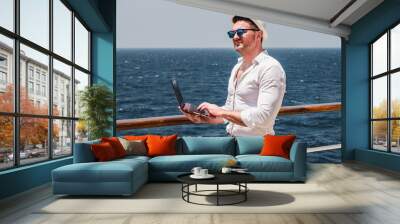 Fashionable man in sunglasses and a sun hat, working on a red laptop on the empty deck of a cruise liner against the background of sea waves. View from the back. Concept of leisure and travel
 Wall mural