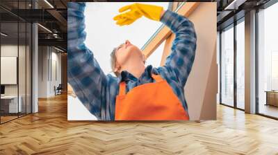 woman in blue checkered shirt and orange apron holding window cleaner sprayer and cloth cleaning house and washing windows Wall mural