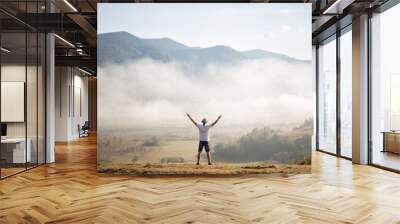 Happy traveler man raising hands up on the top of the world above mountains in white clouds. Hipster guy enjoy amazing atmospheric moment. travel and wanderlust concept Wall mural