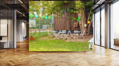 Place for children birthday party front or back yard with hanging decorative multi-colored balloons, white tables and chairs, bench near a wooden house in green autumn grass Wall mural