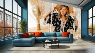 Cozy home atmosphere.  Lovely stylish blond woman in boho dress posing in stylish bedroom. Ladysitting on hanging swing . . Wall mural