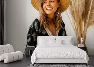 Laughing blond woman in straw hat and boho dress  posing in studio over white background  with pampas grass decor.  Pillows with macrame ornament. Summer trends. Wall mural