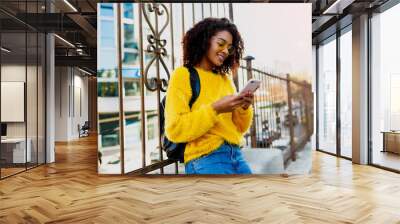 Outdoor image of young attractive black woman with stylish Afro hairs  using mobile phone and sitting on  bridge  in modern district. Yellow glasses and pullover. Wall mural