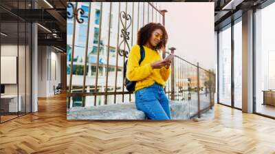 Outdoor image of young attractive black woman with stylish Afro hairs  using mobile phone and sitting on  bridge  in modern district. Yellow glasses and pullover. Wall mural