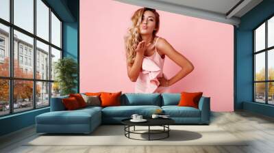 seductive woman in stylish  pink party dress send kiss to camera and flirting   on  rose  wall . Blond  healthy wavy hairs. Fashion portrait. Wall mural