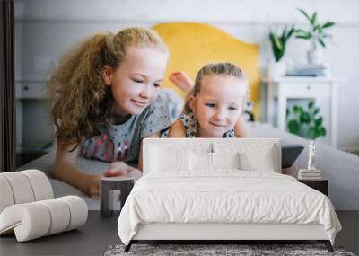 Two beautiful little sisters lying in the bed and look at the screen of a tablet, smart kids using smart technology Wall mural