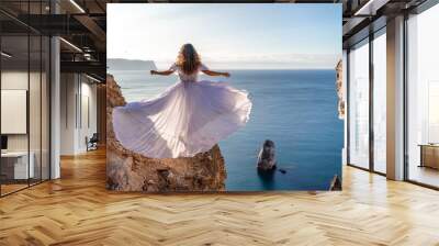 A woman in a white dress stands on a rocky cliff overlooking the ocean. The scene is serene and peaceful, with the woman's dress billowing in the wind. The ocean is calm and blue. Wall mural