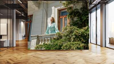 A woman on a balcony in a long blue dress stands near a low granite stone fence against the background of an old stone house Wall mural