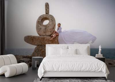 A woman sits on a stone sculpture made of large stones. She is dressed in a white long dress, against the backdrop of the sea and sky. The dress develops in the wind. Wall mural