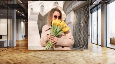 A woman wearing a green dress and holding a bouquet of yellow flowers. She is standing on a white staircase in front of a building. Wall mural