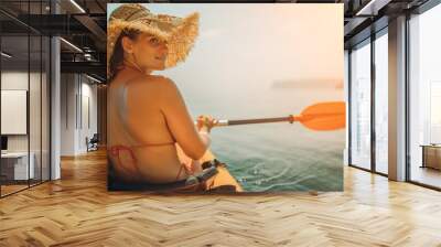 A woman wearing a straw hat is paddling a canoe on a sunny day. Scene is relaxed and carefree, as the woman enjoys her time on the water. Wall mural