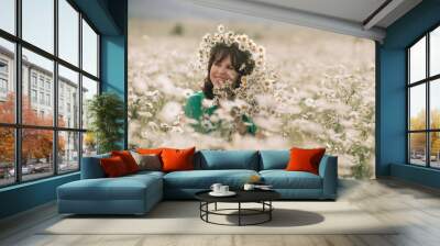 Happy woman in a field of daisies with a wreath of wildflowers on her head. woman in a green dress in a field of white flowers. Charming woman with a bouquet of daisies, tender summer photo Wall mural