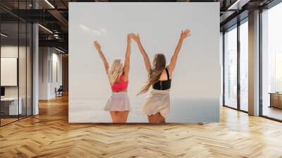 Two women are standing on a beach, one wearing a pink top and the other wearing a black top. They are both smiling and raising their arms in the air, as if they are celebrating something. Wall mural