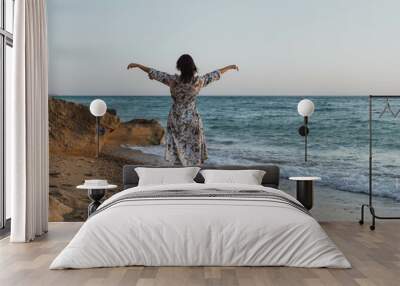 Woman Beach Ocean - A woman wearing a floral dress stands on a beach with her arms outstretched, facing the ocean at sunset. Wall mural