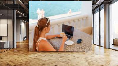 Woman coffee cafe laptop sea. Female freelancer using laptop on coffee break in modern cafe with sea view in the morning Wall mural