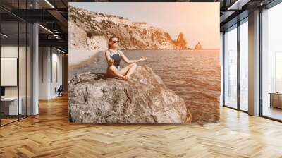 Yoga on the beach. A happy woman meditating in a yoga pose on the beach, surrounded by the ocean and rock mountains, promoting a healthy lifestyle outdoors in nature, and inspiring fitness concept. Wall mural