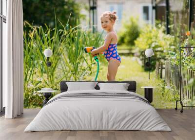 Charming baby girl waters plants with a hose in the garden in the backyard of the house on a sunny summer day Wall mural