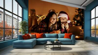 Two cute sisters use a laptop at home by the Christmas tree on Christmas Eve, watch a movie or send Christmas greetings. Winter evening at home with family and children Wall mural