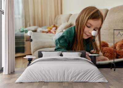 A cute little girl enjoy playing games on her tablet while lying on her stomach on a sofa Wall mural