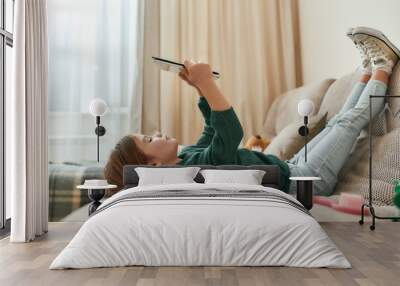 A small cute girl playing games on a tablet lying on a sofa with her feet up and gumshoes on Wall mural