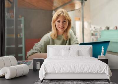 A smiling and well-dressed blondy making notes to a flipbook while sitting at a table in front of a laptop looking into a camera Wall mural