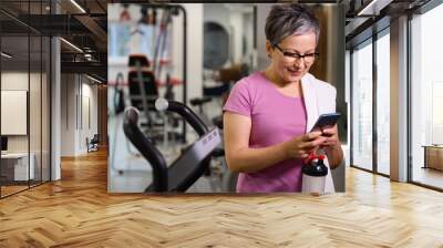 Adult smiling caucasian woman wearing sportswear using smartphone in gym Wall mural