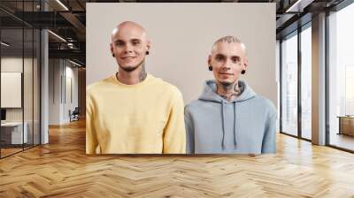 Beat friends. Portrait of young happy caucasian twin brothers with tattoos and piercings smiling at camera while posing together in studio, standing isolated over beige background Wall mural