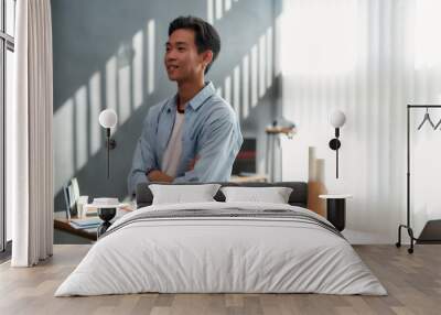 Confident designer. Side view of a young asian man in casual wear with crossed arms looking away and smiling while standing in the modern office Wall mural