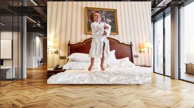 Discover happiness here. Cheerful boy in white bathrobe is having fun, jumping on the white bed in a hotel room. The kid laughs and jumps Wall mural