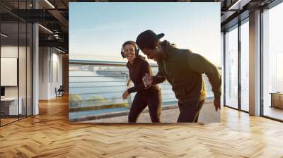 Enjoying run. Side view of happy young African couple, man and woman in headphones laughing while running on the bridge in the early morning Wall mural