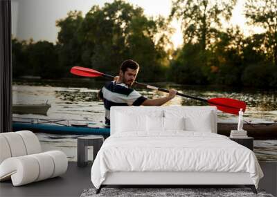 Enthusiastic young caucasian guy holding a paddle while boating on a lake surrounded by nature Wall mural