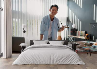 Happy at work. Cheerful asian man holding eyeglasses and smiling at camera while leaning on a table with a lot of creative stuff on it. Young designer in casual wear working in the modern office Wall mural