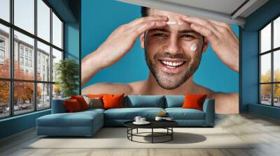 Happy brunette guy smiling at camera while applying white cream on his face, posing isolated over blue background Wall mural