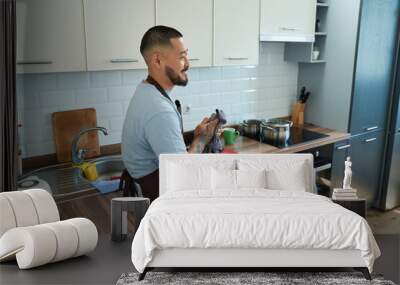 Joyful man stands kitchen cabinet and wipes hands with kitchen towel Wall mural