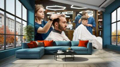 Man visiting barbershop. Side view of a professional barber girl making new modern haircut for a young bearded man Wall mural