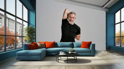 Middle aged muscular man in black sportswear looking at camera while warming up his body before workout in studio over grey background Wall mural